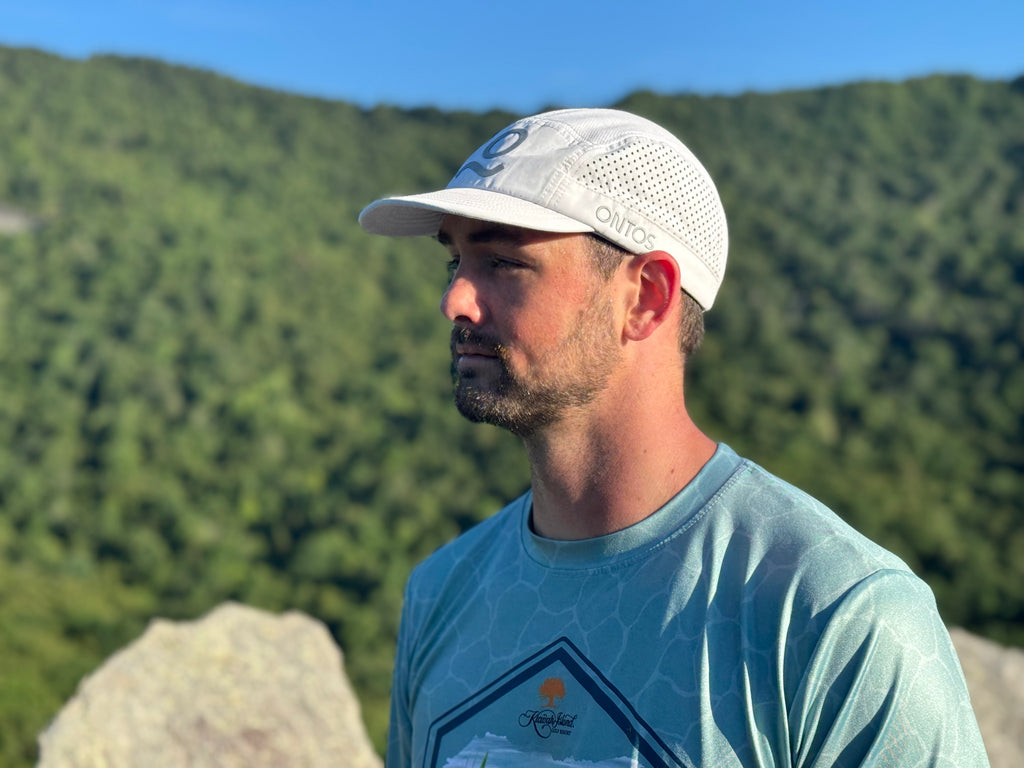 A man wearing a white outdoor mesh cap and a light blue patterned t-shirt, standing outdoors with a forested mountain in the background.
