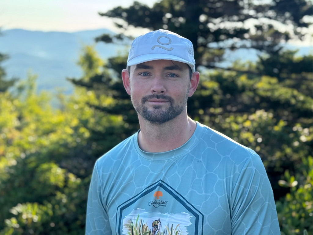 A man wearing a light blue patterned t-shirt and a matching light blue cap stands outdoors in front of a blurred mountain and forest background.